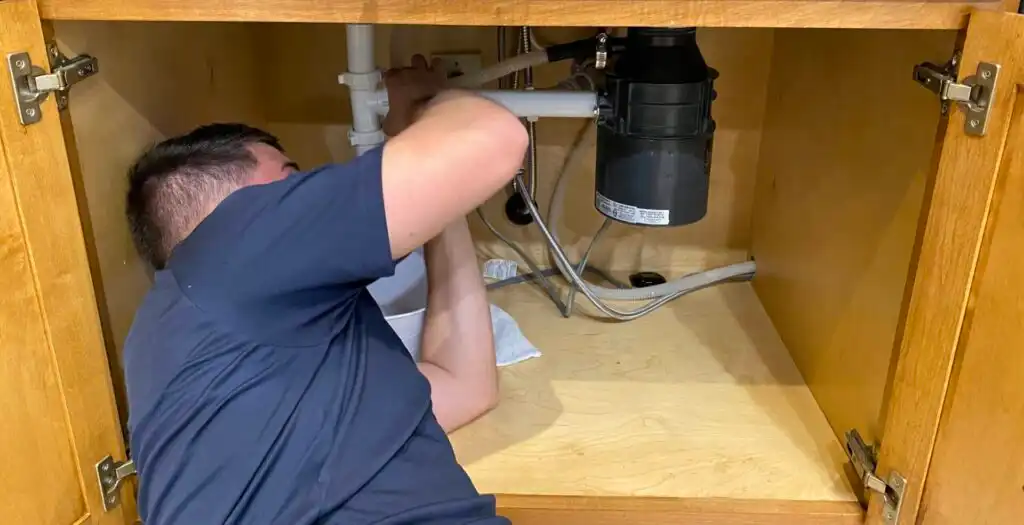 Technician working under a kitchen sink, installing or servicing plumbing components including a black drain assembly and white PVC pipes.