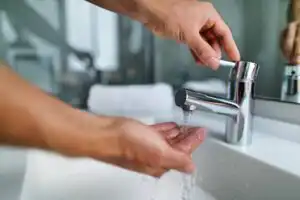 Person turning off a chrome bathroom faucet while washing hands under running water at a white sink.