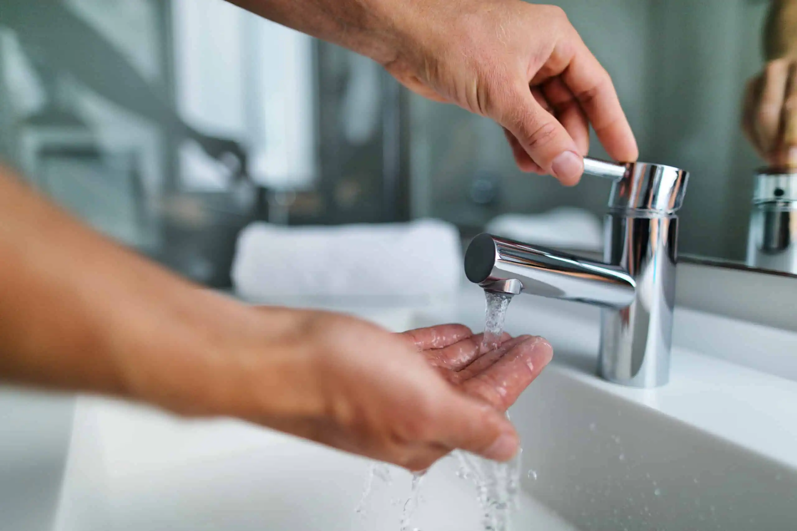 Person turning off a chrome bathroom faucet while washing hands under running water at a white sink.