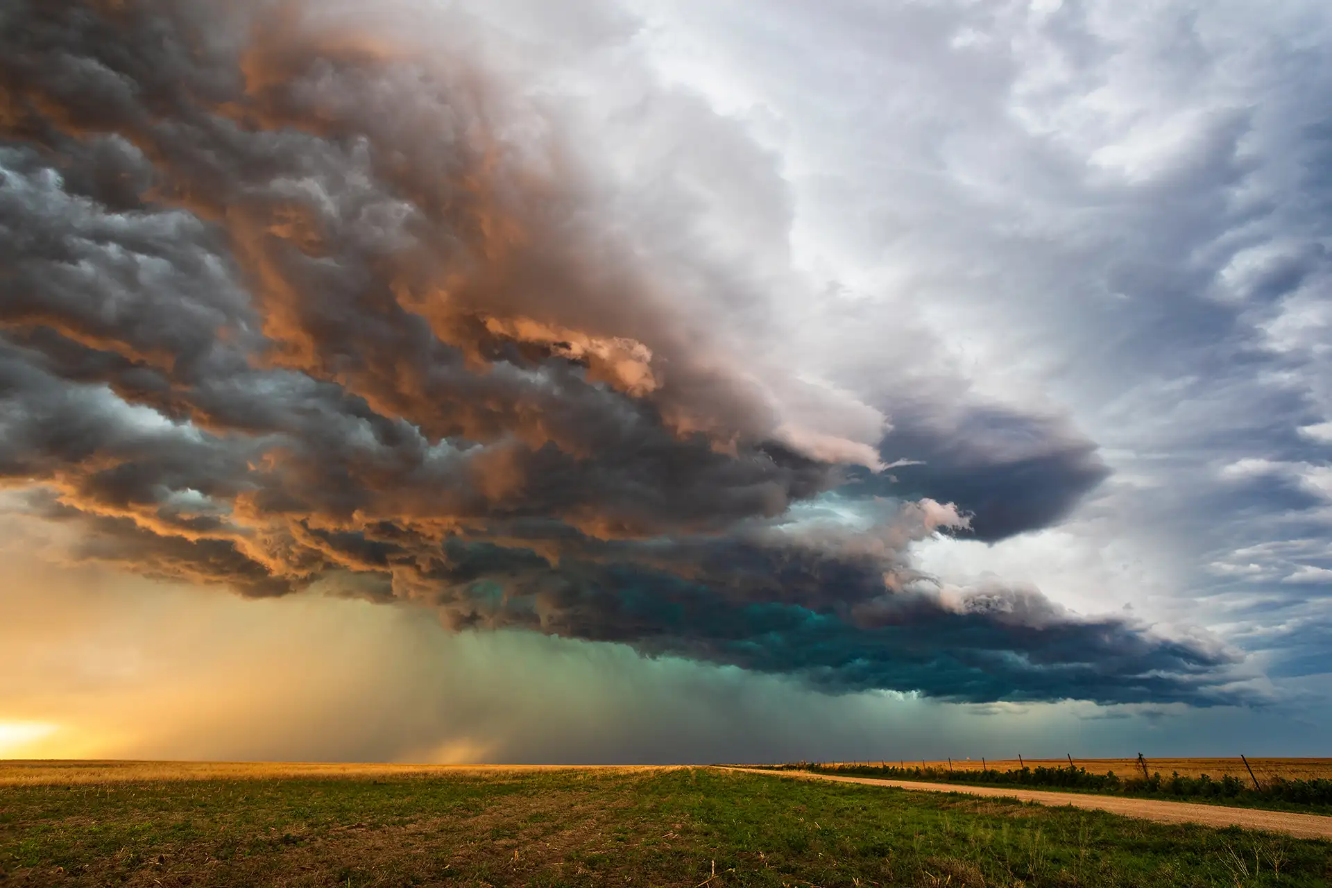 Massive supercell thunderstorm cloud formation with golden and teal colors looming over flat grassland with dirt road during sunset.