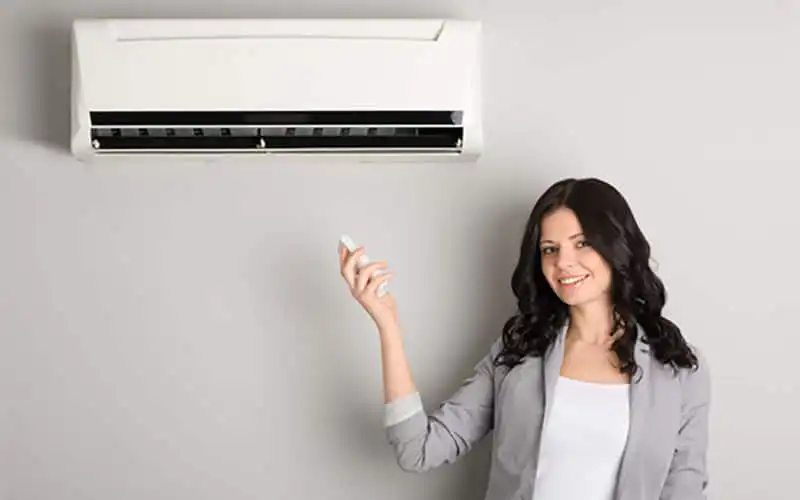 Woman in business casual attire holding remote control while standing below a wall-mounted ductless mini split air conditioning unit.