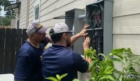 Two technicians in navy uniforms installing or servicing an electrical panel on the exterior of a residential home.