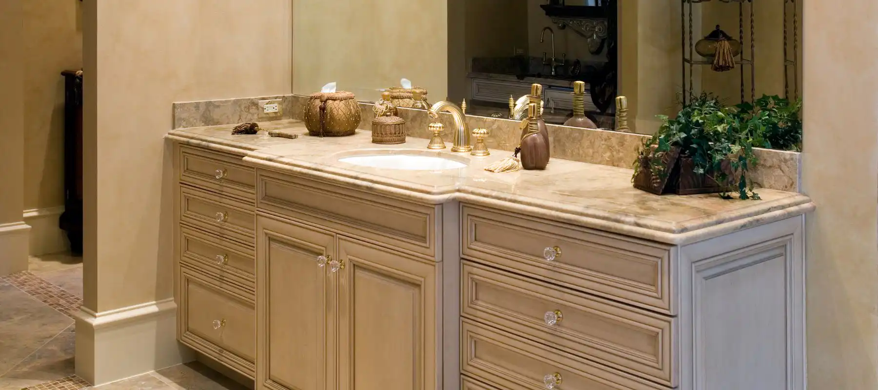Elegant bathroom vanity featuring dual marble sinks, brass fixtures, cream cabinetry with decorative hardware, and decorative accessories.