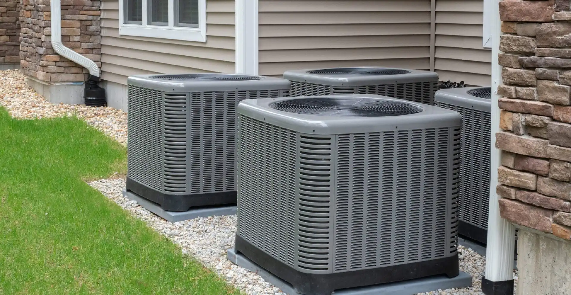 Three gray HVAC condenser units installed on gravel beside a brick home with siding and white trim.