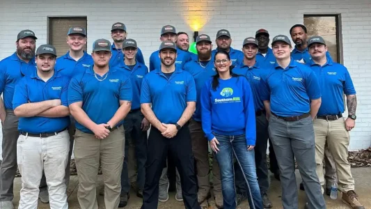 Professional team of HVAC, plumbing, and electrical technicians wearing blue uniforms and caps standing together in front of white brick building.